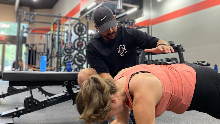 Sami, entraîneur chez Studio Fit U qui démontre à sa cliente Maria une planche pour l'activation de ses abdominaux.