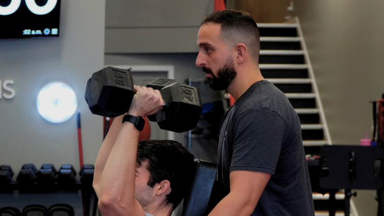 Sami assisting a client with a dumbbell shoulder press at the gym.