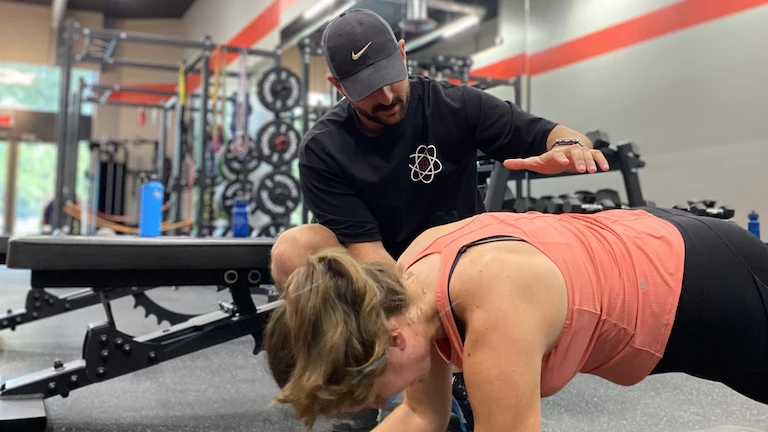 Sami, entraîneur chez Studio Fit U qui démontre à sa cliente Maria une planche pour l'activation de ses abdominaux.